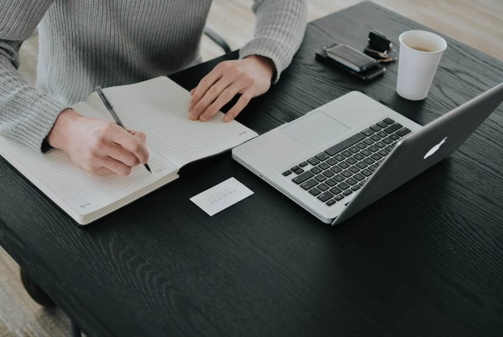Person writing in a notebook at a desk with a laptop, showcasing industry-specific business CRM planning
